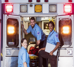 A multi-ethnic group of three paramedics at the rear of an ambulance, ready to climb in through the open doors. The two women their male colleague are smiling at the camera.