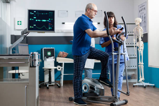 Old patient doing physical exercise and measuring pulse on heart rate monitor for muscle recovery. Senior person using electrical stationary bike for rehabilitation with nurse assisting