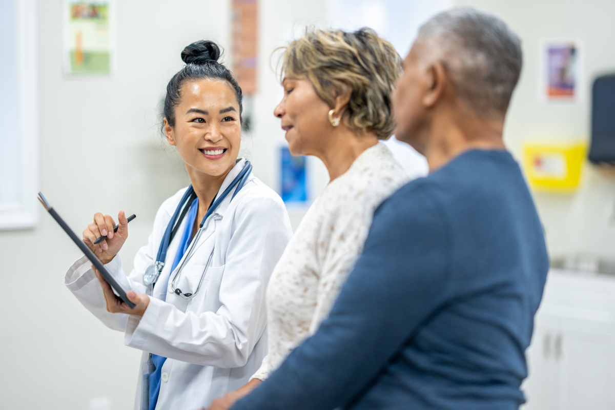 A senior patient, of Hispanic decent, sits up on an exam table with her husband close by as she receives her recent tests results. The couple appears happy as they receive good news from the doctor