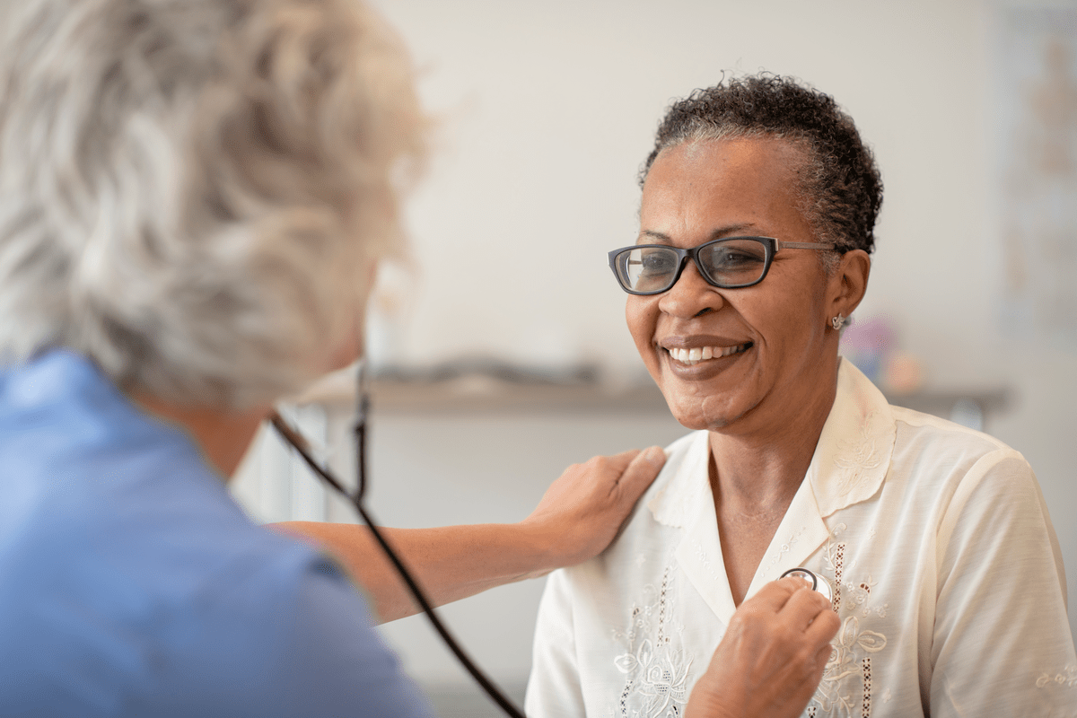 A senior black women is having her heart checked by a female doctor. The patient is wearing glasses and smiling at the doctor. The doctor is wearing scrubs and checking the patients heart rate with a stethoscope.