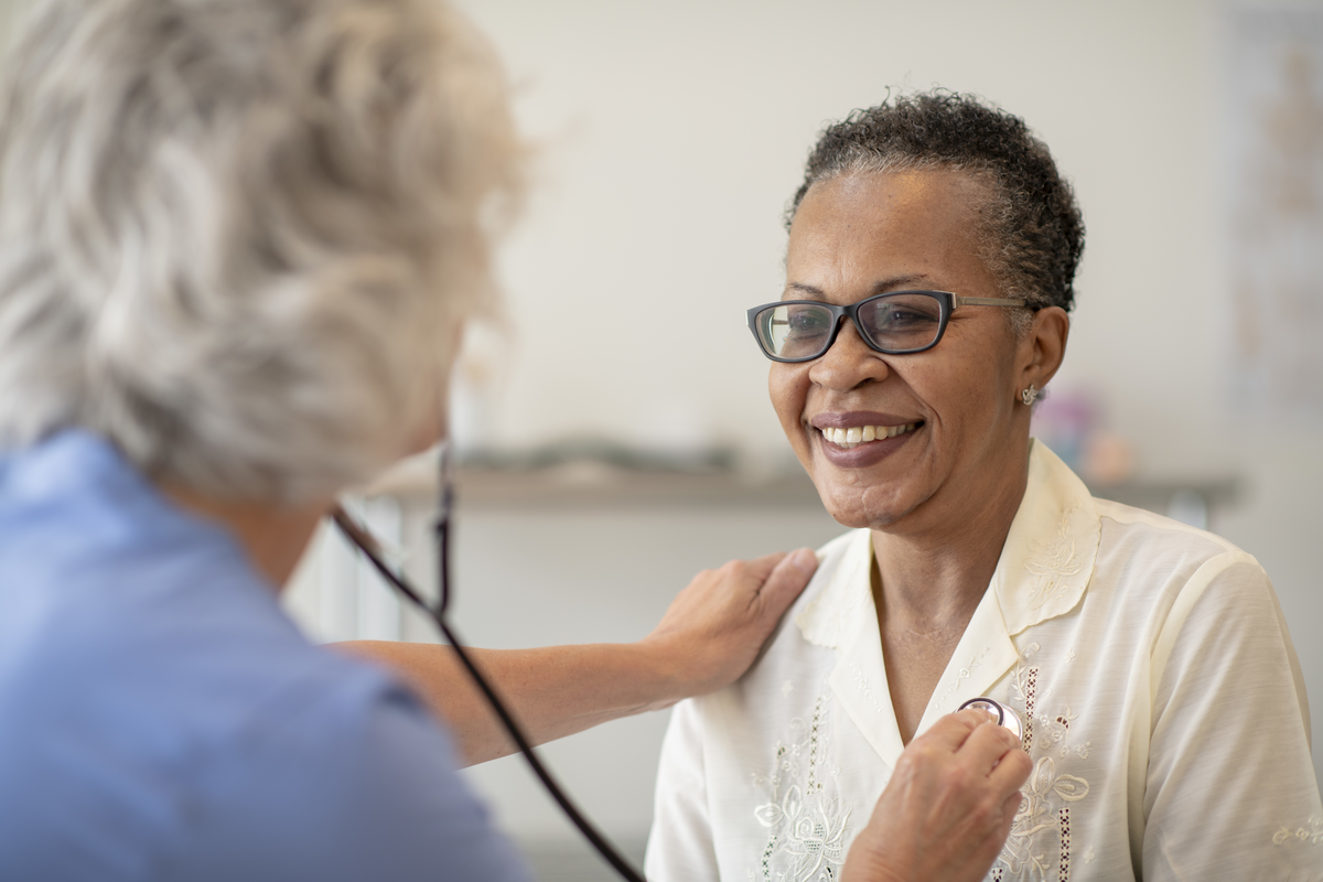 A senior black women is having her heart checked by a female doctor. The patient is wearing glasses and smiling at the doctor. The doctor is wearing scrubs and checking the patients heart rate with a stethoscope.