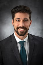 Professional headshot of a smiling bearded man in a dark suit, white shirt, and teal tie against a softly lit gray background.
