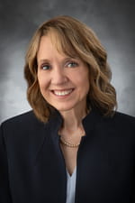Professional headshot of a smiling woman with wavy brown hair wearing a black blazer and pearl necklace against a gray background.