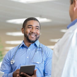 A smiling doctor with a stethoscope around his neck holds a tablet, engaged in conversation with another medical professional, conveying a friendly atmosphere.