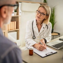 A female doctor with glasses consults an older male patient. She writes on a clipboard, conveying professionalism and attentiveness in a cozy office.