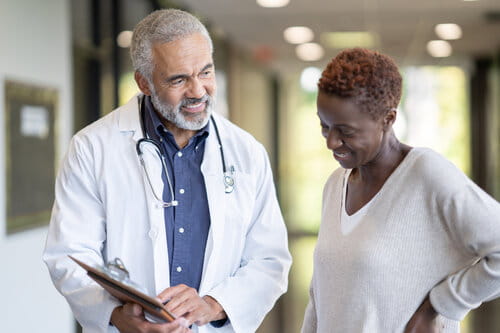 A doctor with gray hair and a stethoscope smiles while holding a clipboard, talking to a woman in a hallway. They appear to be having a friendly conversation.