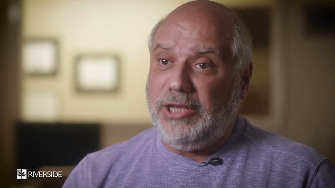 A man with a beard speaks in a softly lit room, wearing a lavender top. The expression conveys engagement. The Riverside logo is visible in the corner.