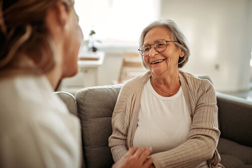 Elderly woman with glasses smiles warmly at another person while sitting on a grey sofa in a well-lit room, conveying a sense of warmth and connection.