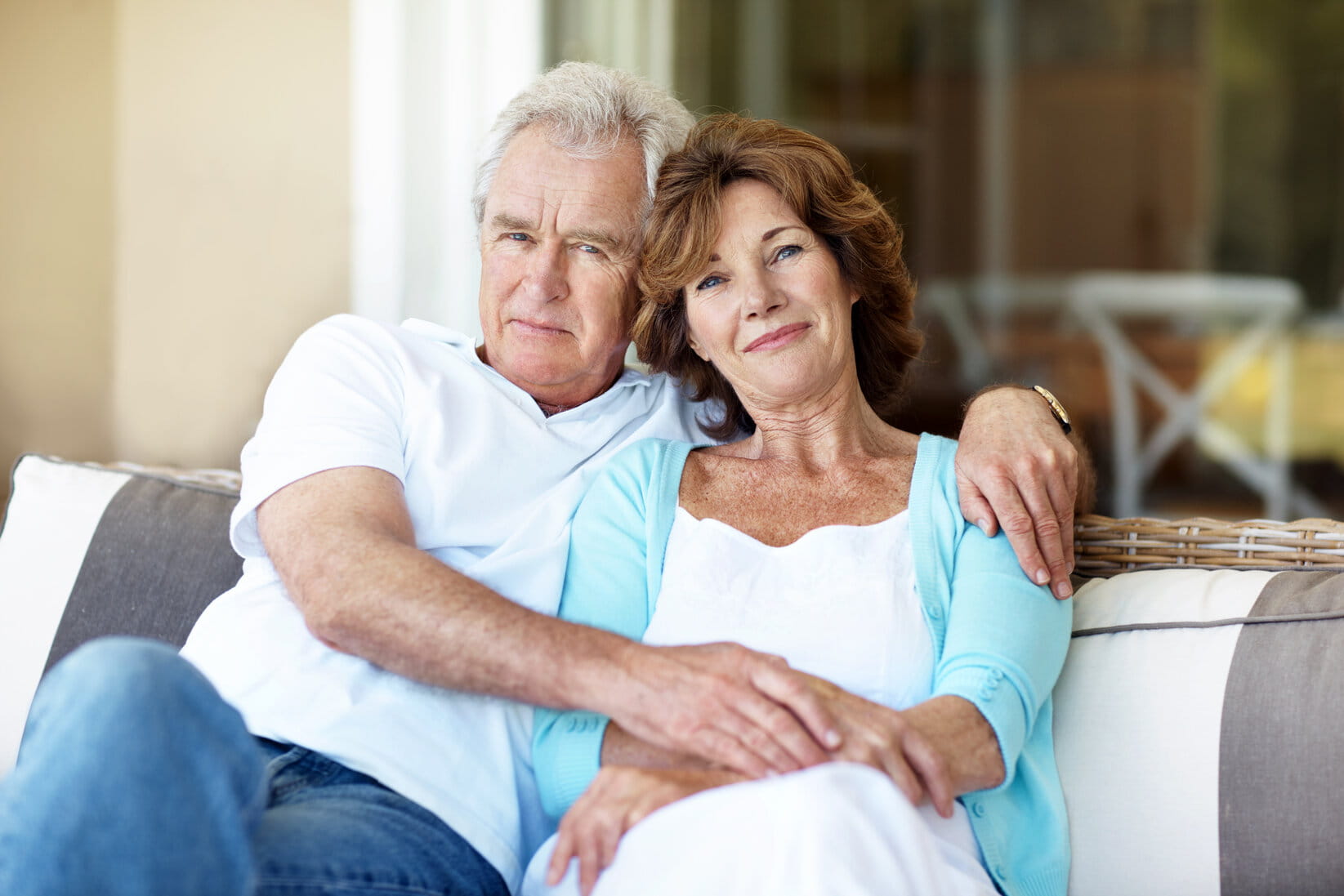 Older adults sitting together on a sofa in a home setting, relaxing with arms around each other.