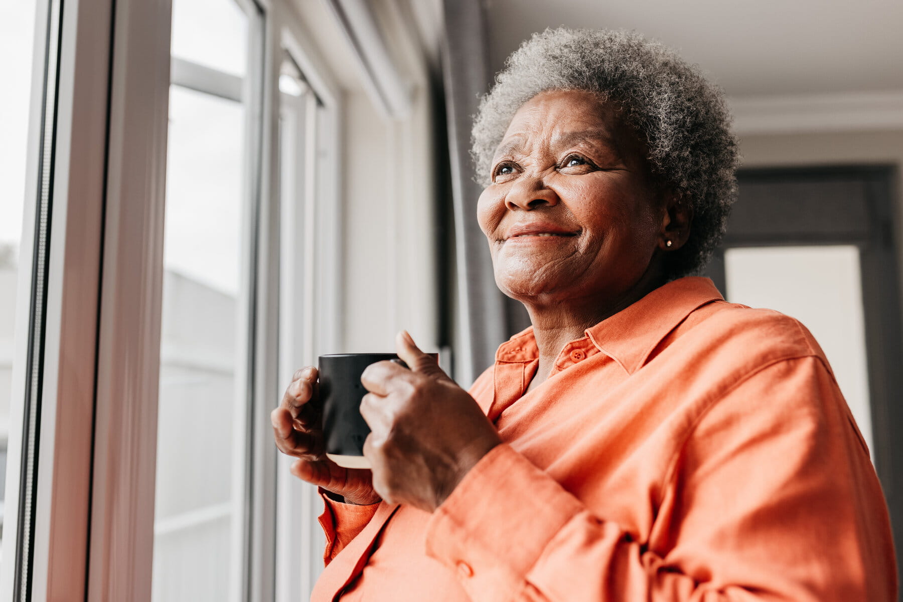 A smiling elderly woman in an orange shirt holds a mug, gazing out a bright window. She appears content and reflective in a cozy indoor setting.