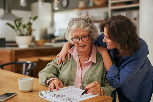 A smiling woman embraces an elderly woman doing a crossword at a kitchen table. The scene is warm and cozy, with a mug nearby.