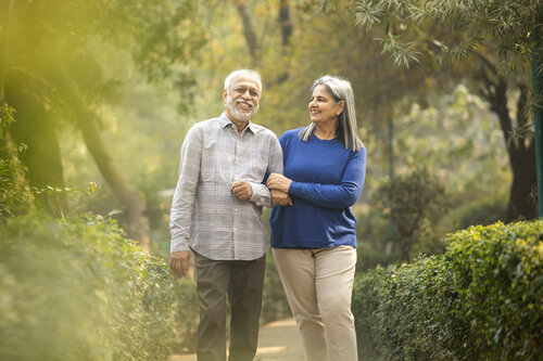 An elderly couple walks arm in arm through a lush, green park. They are smiling and enjoying a sunny day, creating a serene and joyful atmosphere.