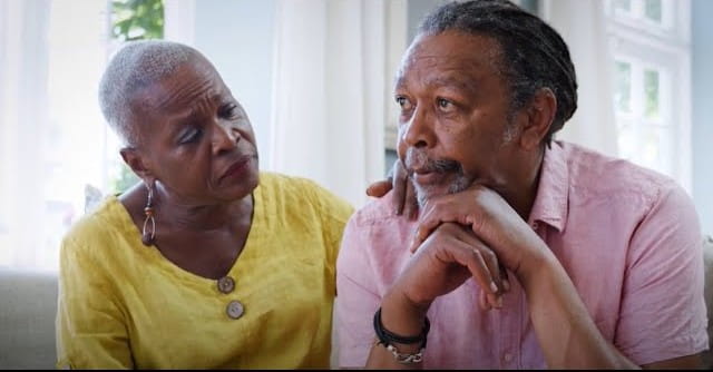 An elderly woman in a yellow blouse gently comforts a thoughtful, concerned man in a pink shirt, creating a supportive and caring atmosphere.