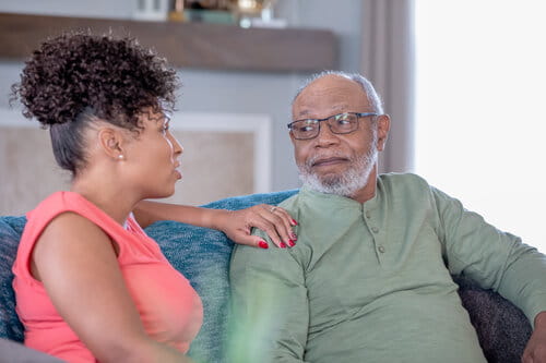 An elderly man in glasses and a young woman with curly hair are sitting on a sofa, engaged in a warm, thoughtful conversation. The setting is cozy and homey.