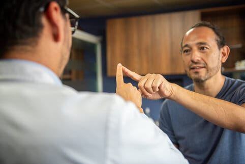 A man in a blue shirt and a person in glasses point at each other's fingers in synchronicity, appearing focused, indoors with wooden cabinets in the background.