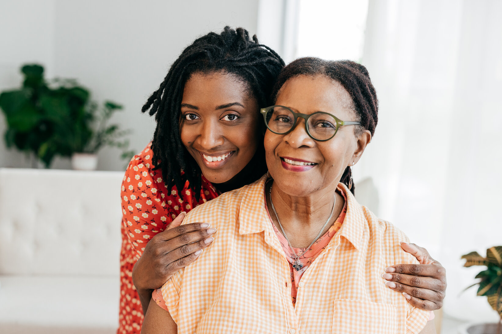 A young woman in a red shirt embraces an older woman in glasses and a yellow checkered blouse, both smiling warmly in a bright, cozy room.