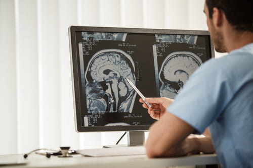 A doctor in blue scrubs examines brain scans on a computer monitor, pointing with a pen. The setting is clinical, focused, and analytical.