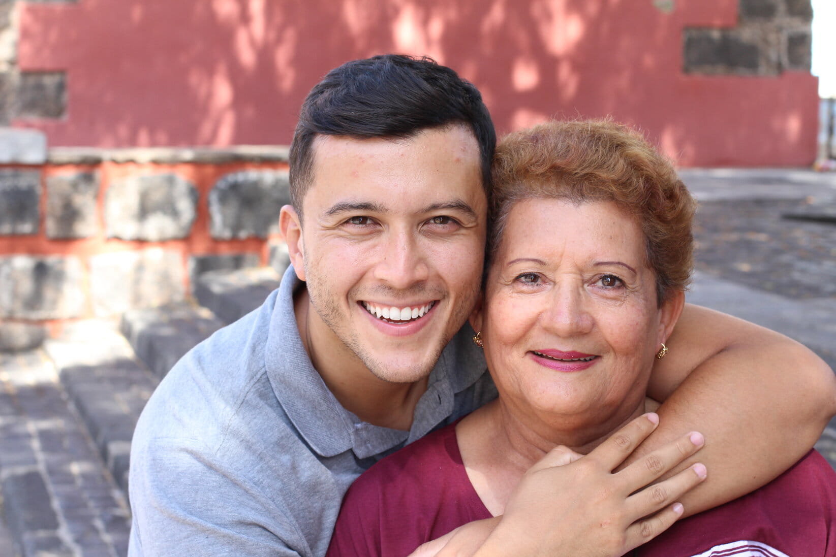 A smiling young man hugs an older woman affectionately. They are outdoors with a red wall and stone steps behind them, conveying warmth and joy.