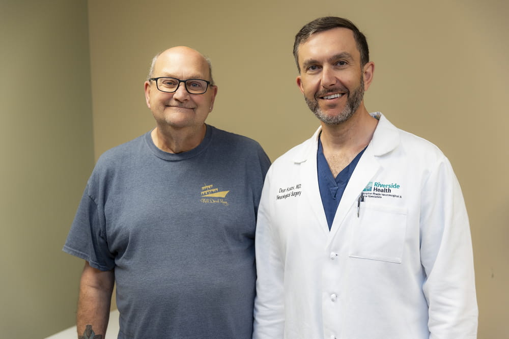Older male patient standing beside a surgeon in a white coat in an exam room, both smiling at the camera.