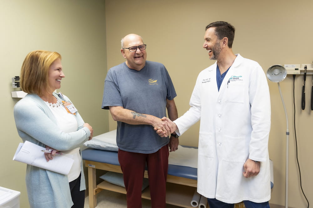 Older male patient shaking hands with a surgeon in an exam room while a staff member stands nearby smiling.