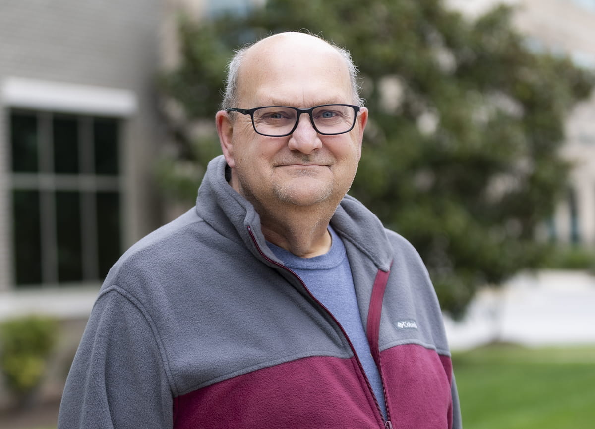 Portrait of an older man wearing glasses and a gray-and-maroon fleece jacket, standing outdoors in front of a building and greenery.