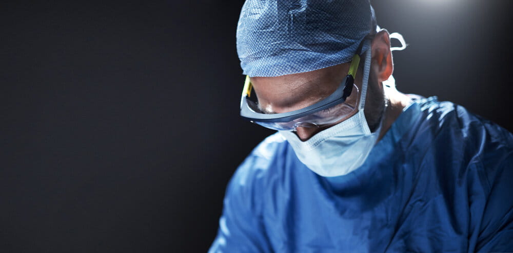 A focused surgeon in blue scrubs, cap, mask, and goggles operates under intense light in a dimly lit room, conveying precision and concentration.