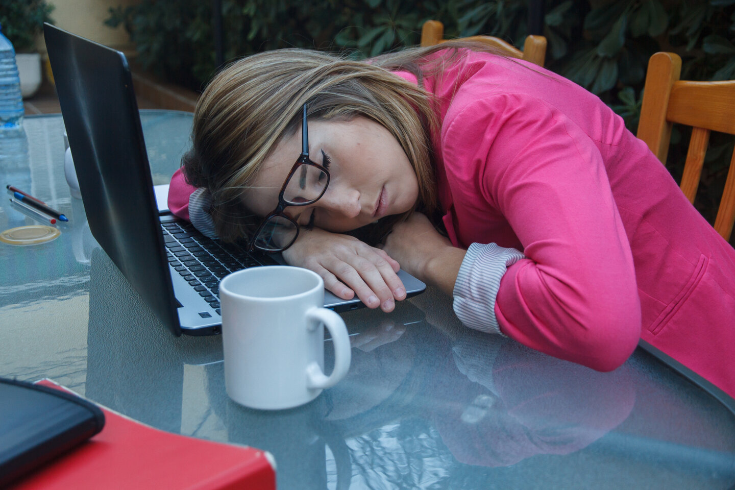 A woman in glasses and a pink blazer is asleep on a laptop at a glass table. Nearby is a white mug, creating a tired and overworked atmosphere.