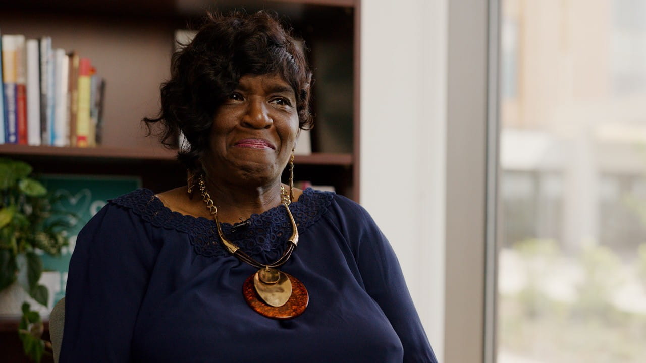 Portrait of an older Black woman in a navy blouse and layered gold necklace, seated before a bookshelf, smiling warmly in soft light.