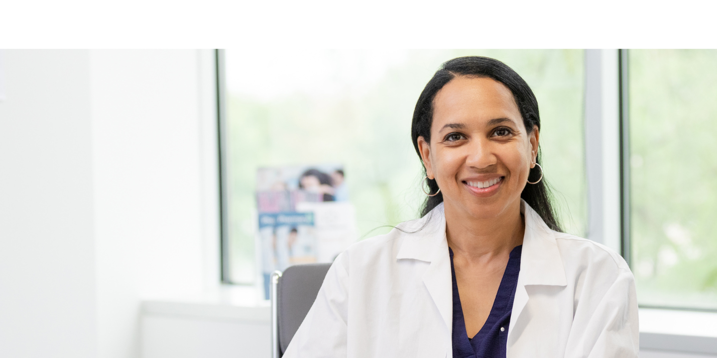 Looking at the camera, the mature adult female doctor sits with her hands clasped in front of her on the desk.