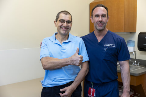 Two men are smiling in a doctor's office. One is wearing a light blue polo, giving a thumbs up, while the other is in navy blue scrubs, standing confidently.