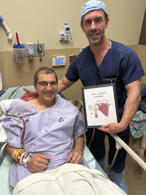 A smiling patient sits in a hospital bed, wearing a gown with medical wristbands. A doctor in scrubs stands beside them, holding a diagram of a shoulder. The scene is positive and reassuring.