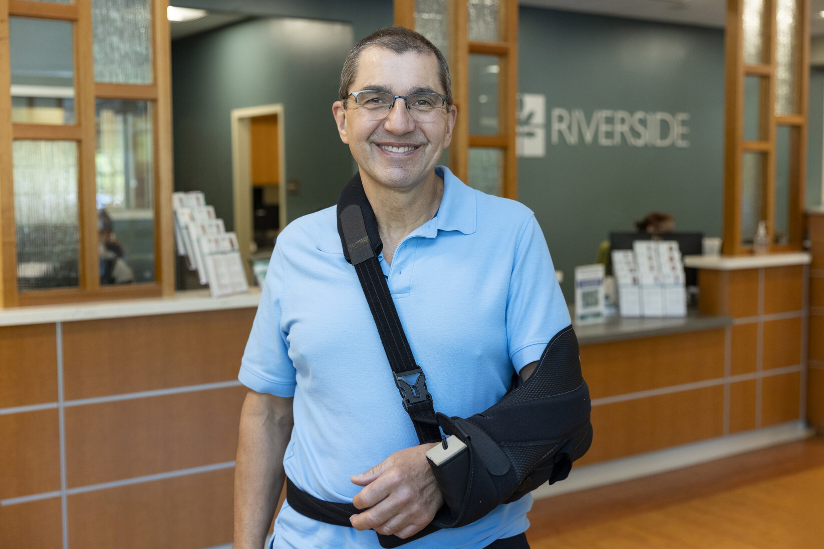Smiling man in blue shirt with arm sling stands in a medical office lobby, possibly named Riverside. The mood is positive and welcoming.
