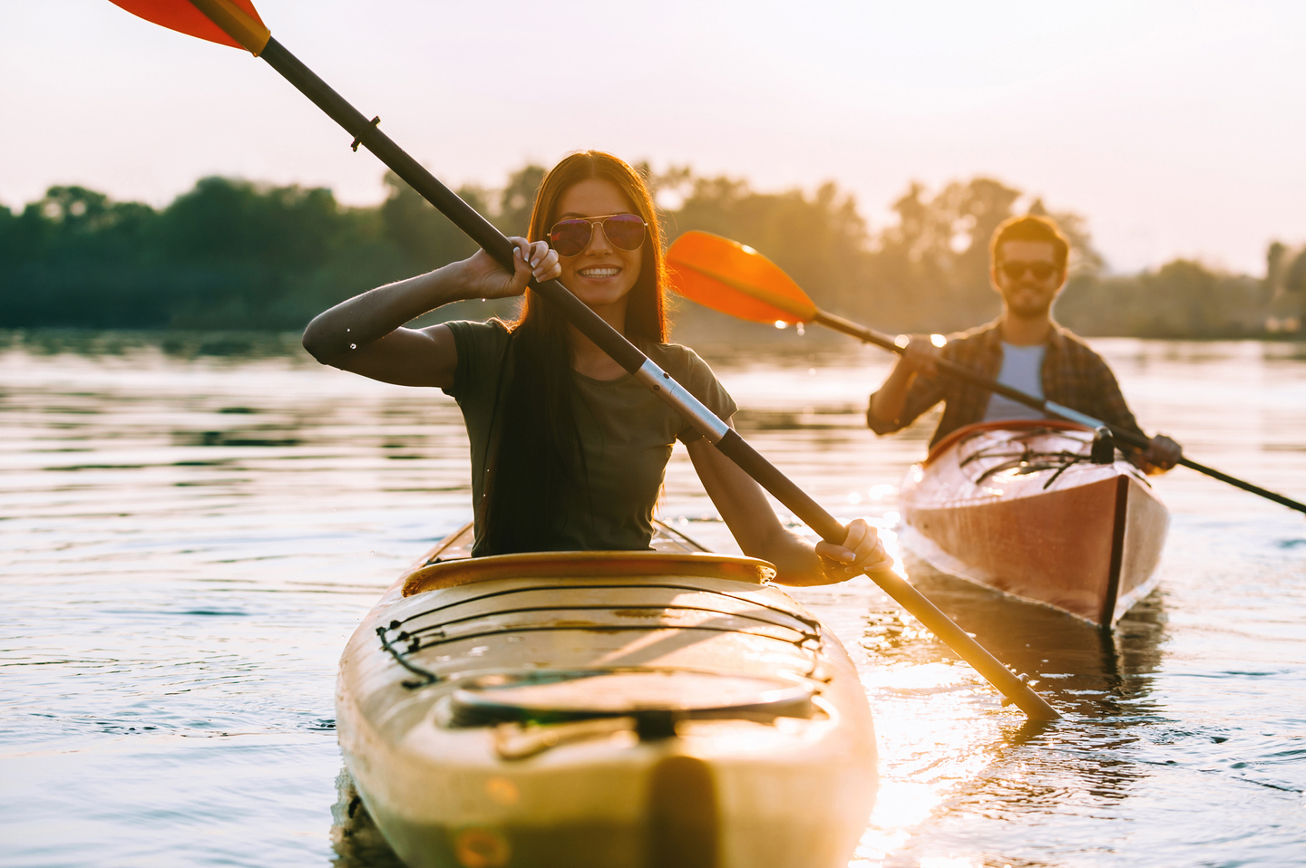 Two people kayaking on a calm river at sunset, smiling and paddling together.