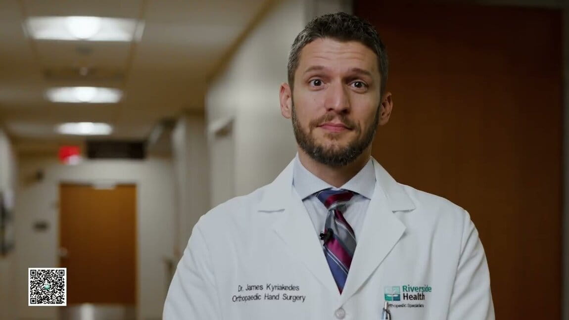 A man in a white coat stands in a hallway with warm lighting, conveying professionalism. The coat reads "Orthopaedic Hand Surgery," and he appears approachable.