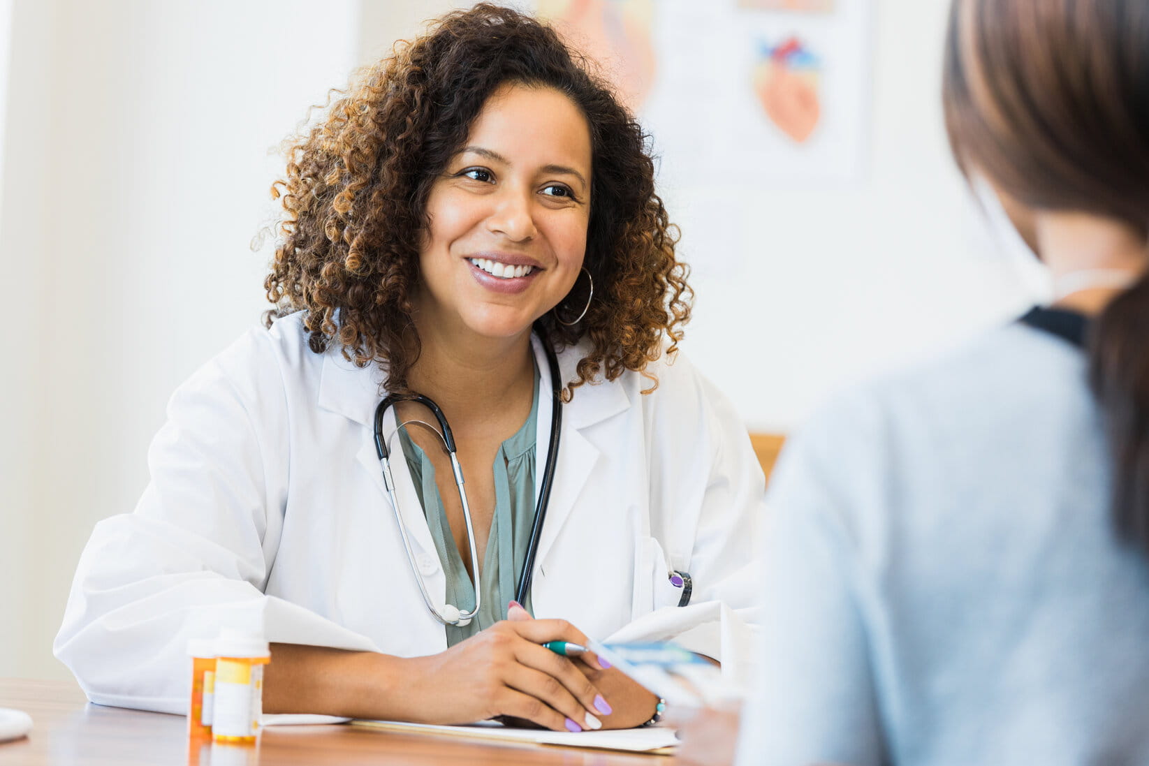 A smiling doctor in a white coat listens attentively to a patient, shown from the back, in a bright exam room. The atmosphere is warm and supportive.