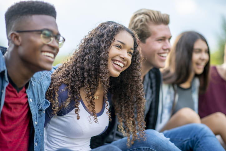 A group of multi-ethnic students are sitting outside in the grass together, with their arms around one another. They are sharing a laugh and enjoying each others company