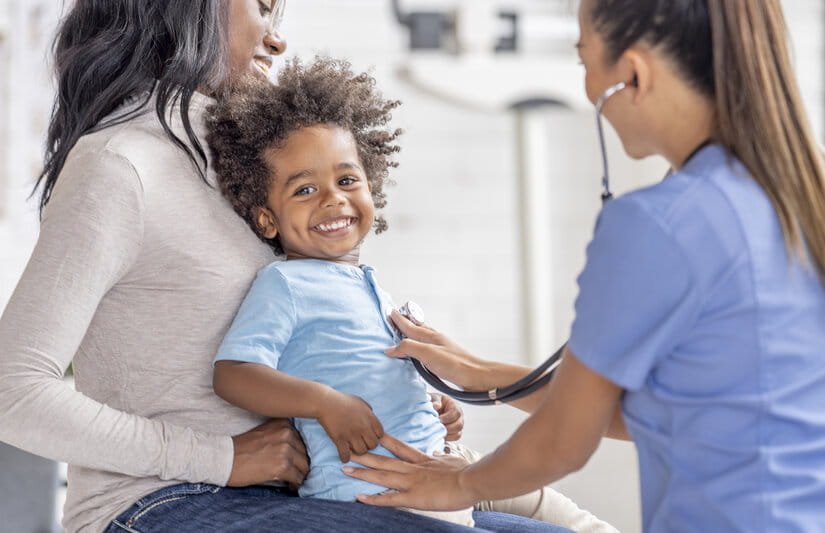 A young mother of African decent sits up on an exam table with her son on her lap as they meet with the doctor. They are both dressed casually and he doctor is wearing blue scrubs as she checks out the young boy.