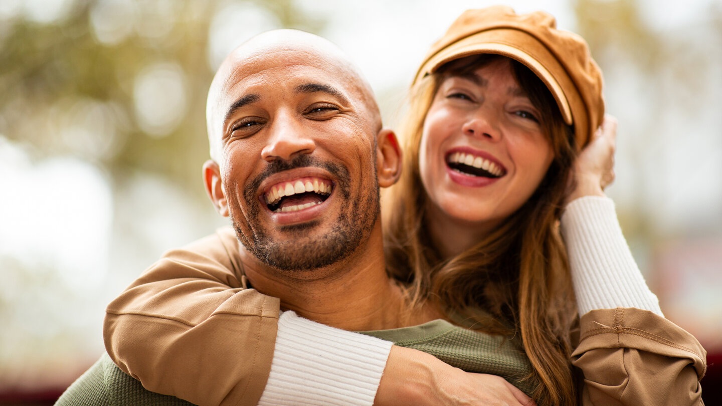 A joyful man and woman share a warm embrace, both smiling widely. The woman wears a tan hat, and an outdoor setting with blurred greenery is in the background.