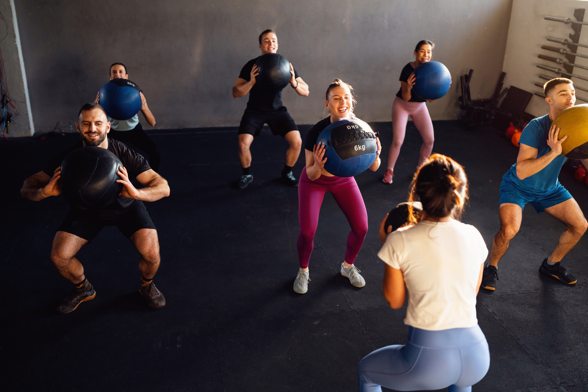 A group of young and active female and male Caucasian people with female cross trainer, exercise with weight balls, during a group workout at the gym