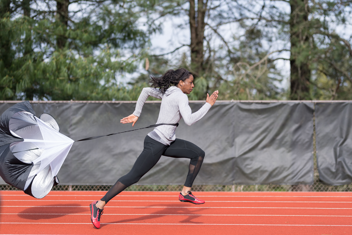 An African American athlete runs sprint drills on a stadium track. She is running with a parachute attached to her for resistance training.