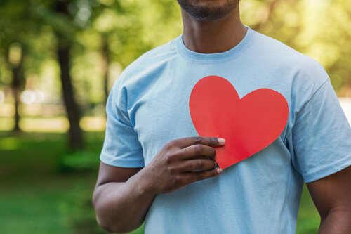 A person in a light blue T-shirt holds a large red paper heart against their chest. They stand outdoors with blurred green trees in the background, conveying warmth.