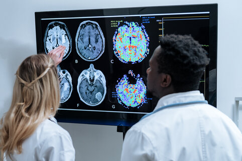 Two medical professionals examine colorful brain scans on a large monitor. The scene is analytical and focused, highlighting diagnostic imagery.