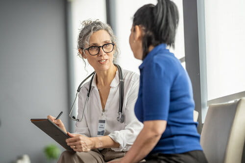 A healthcare professional with glasses and a stethoscope smiles while talking to a seated woman in a blue shirt, conveying a supportive atmosphere.