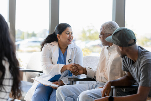 A friendly doctor in a white coat smiles and shakes hands with an elderly man in a waiting room. Another person watches, creating a warm atmosphere.