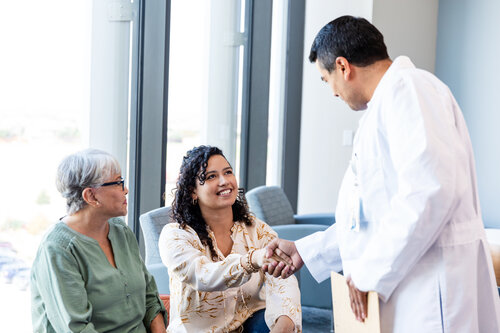 A doctor in a white coat warmly shakes hands with a smiling woman, seated beside an older woman, in a bright room, conveying trust and care.