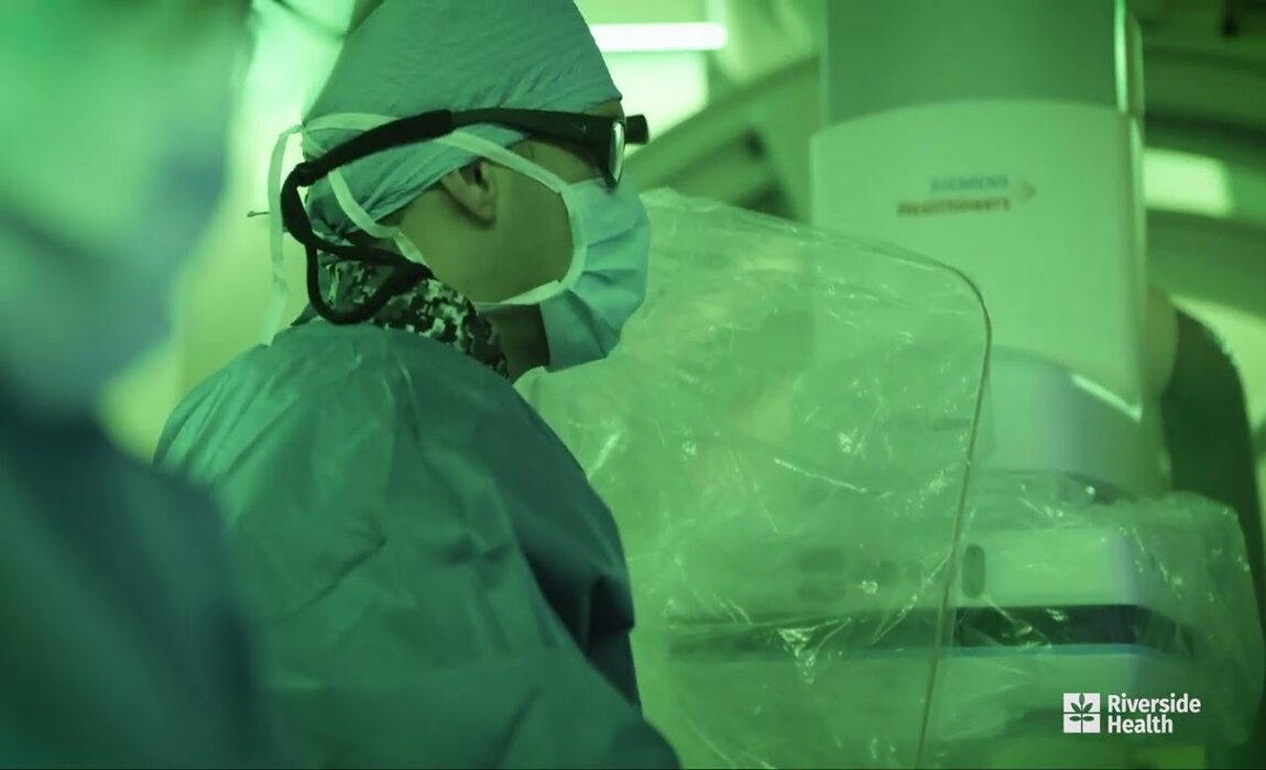 A surgeon in green scrubs and mask operates under green lighting, focused on medical equipment. The mood is serious, with a Riverside Health logo visible.