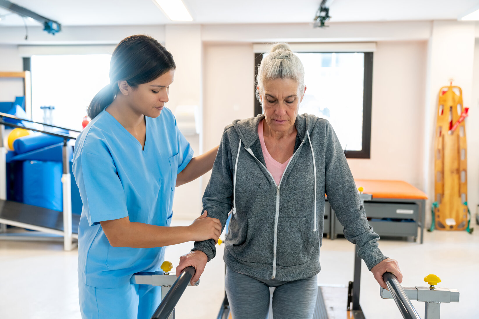 A healthcare worker in blue scrubs supports an older woman using parallel bars for physical therapy in a bright, well-equipped rehab facility.