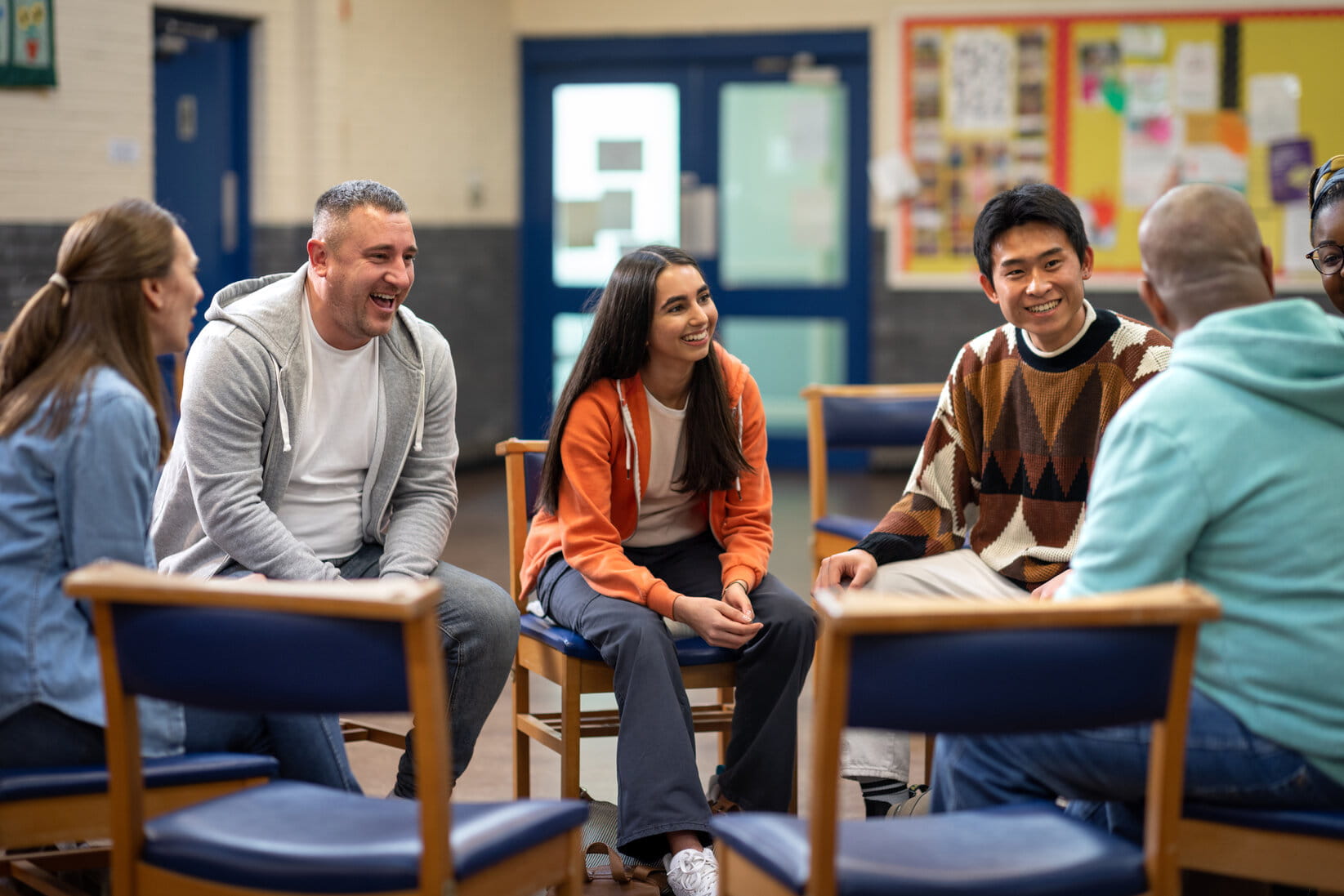 A diverse group of people sit in a circle, smiling and engaging in a lively discussion in a community center setting. The atmosphere is friendly and open.