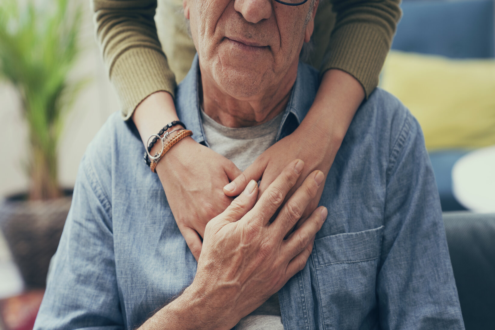 Elderly man wearing glasses and a denim shirt is embraced from behind by a person with crossed hands, conveying warmth and affection.