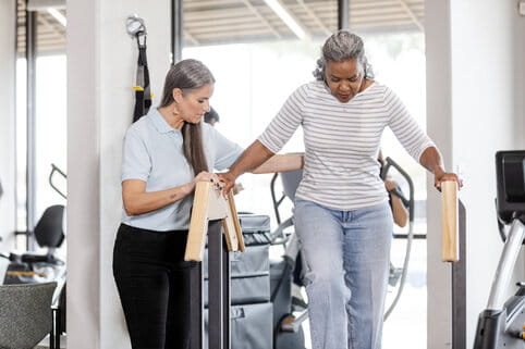 A therapist assists an elderly woman walking between rails in a bright rehabilitation gym. The scene conveys support, focus, and encouragement.